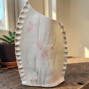 May include: A handmade, off-white ceramic vase with a unique, asymmetrical shape. The vase features a textured, scalloped edge and delicate pink floral designs. The vase is approximately 8 inches tall and is suitable for displaying flowers or as a decorative piece.