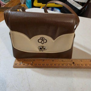 Puede incluir: Bolso de hombro vintage marr&oacute;n y crema con solapa decorativa y cierre plateado. El bolso presenta un emblema de Girl Scout y una correa marr&oacute;n. El bolso mide aproximadamente 20 cm de ancho.