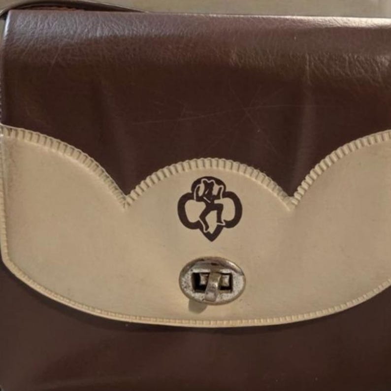 Puede incluir: Un bolso vintage marr&oacute;n y crema con cierre de solapa. El bolso presenta un emblema decorativo y un cierre met&aacute;lico. La solapa crema tiene un borde festoneado. El bolso es probablemente de cuero o material similar.