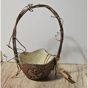 May include: A handcrafted decorative basket with a unique design. The basket features a brown, textured ceramic bowl with a speckled interior and a mosaic-like exterior. A handle made of intertwined twigs arches over the bowl, adding a rustic touch.
