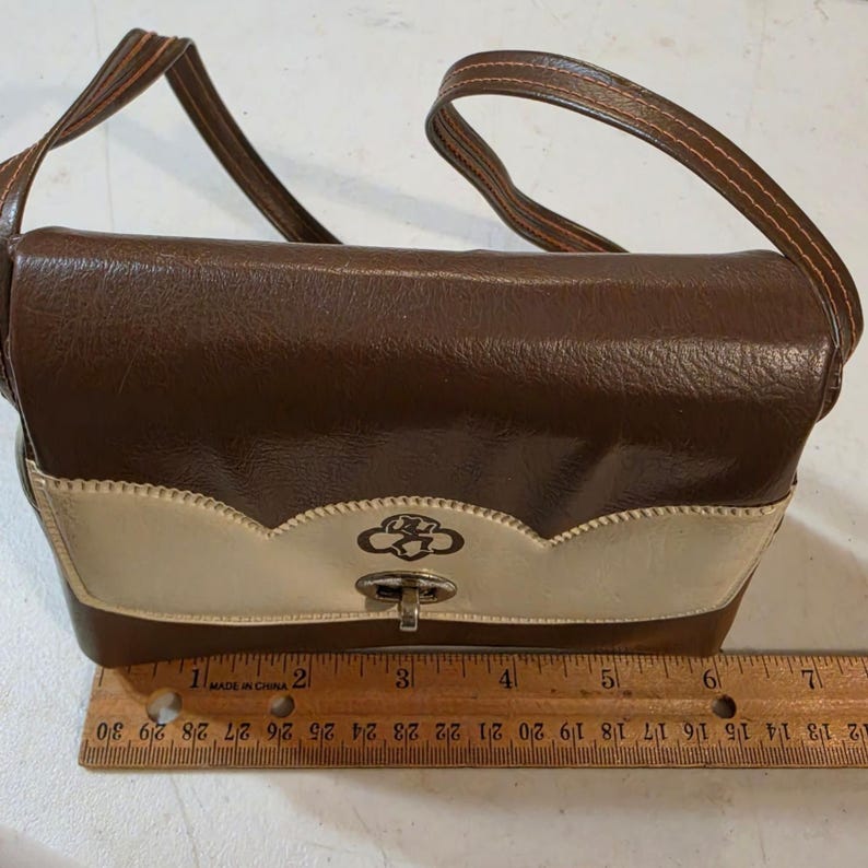 Puede incluir: Un bolso vintage marr&oacute;n y crema con un cierre decorativo y una correa para el hombro. El bolso presenta un dise&ntilde;o festoneado en la parte delantera y un logotipo. La regla junto al bolso muestra medidas en cent&iacute;metros.