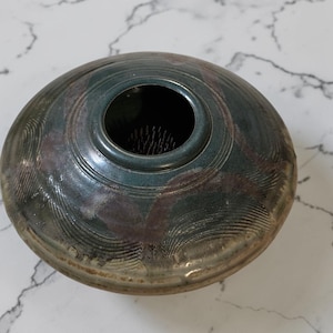 May include: A ceramic flower frog with a dark teal and brown glaze. The round vessel has a central opening surrounded by concentric rings and a textured surface. The frog is designed for arranging flowers.