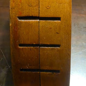 May include: Close-up of a rectangular, wooden object with a warm, brown hue. The wood grain is visible, and there are three horizontal, rectangular cutouts. The object appears to be a component of a larger structure or piece of furniture.