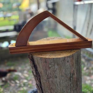 May include: A handcrafted wooden display stand with a curved top and angled support. The stand is made of a rich, warm-toned wood and is designed to hold an item. The stand is displayed on a tree stump.