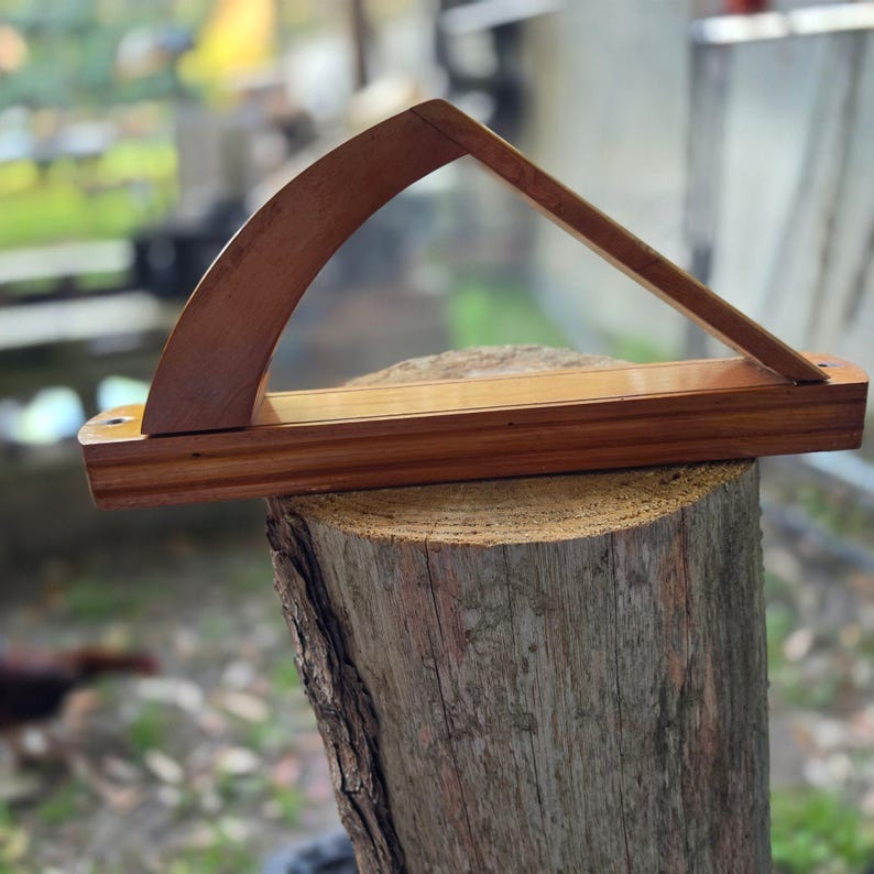 May include: A handcrafted wooden display stand with a curved top and angled support. The stand is made of a rich, warm-toned wood and is designed to hold an item. The stand is displayed on a tree stump.