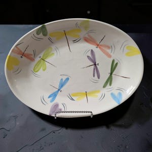 May include: An oval ceramic platter with a white base featuring hand-painted dragonflies in various colors including yellow, green, blue, purple, and orange. Each dragonfly is depicted with small curved lines around the wings to suggest motion.