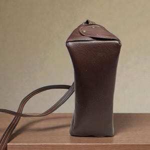 Puede incluir: Un bolso de hombro de cuero marr&oacute;n oscuro con base rectangular y parte superior redondeada. El bolso tiene una correa larga y fina y un cierre de solapa asegurado con dos peque&ntilde;os remaches redondos. El bolso est&aacute; sobre una superficie de madera.