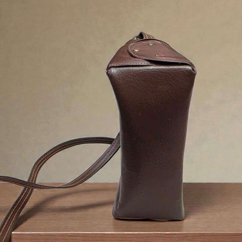 Puede incluir: Un bolso de hombro de cuero marr&oacute;n oscuro con base rectangular y parte superior redondeada. El bolso tiene una correa larga y fina y un cierre de solapa asegurado con dos peque&ntilde;os remaches redondos. El bolso est&aacute; sobre una superficie de madera.