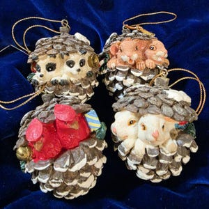 May include: Four decorative Christmas ornaments. Each ornament is shaped like a pinecone and features a different animal: hedgehogs, chipmunks, cardinals, and rabbits. The ornaments have gold hanging strings.