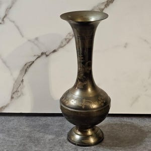 May include: A vintage brass vase with a flared top and a bulbous body. The vase features etched floral designs and a pedestal base. The metal has a dark, aged patina, giving it a classic, antique look.