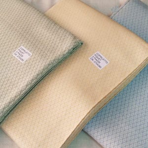 Peut inclure: Trois pièces de tissu pliées en vert clair, beige et bleu clair. Chaque pièce présente un motif en losange et une étiquette blanche avec du texte. Les tissus sont probablement de la literie ou des textiles décoratifs.