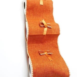 Può includere: Nastro decorativo arancione dall'aspetto strutturato, con tre piccoli fiocchi dorati. Il nastro è arrotolato e presenta un bordo bianco e beige. Ideale per lavori creativi e confezioni regalo.