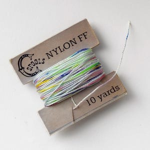 May include: A spool of multi-colored nylon thread, labeled "Spools Nylon FF" and "10 yards." The thread is a mix of white, pink, green, yellow, and blue. The spool is partially unwound, with a length of thread extending from the box.