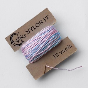 May include: A spool of nylon thread in shades of pink and blue, labeled "SPOOLS NYLON FF" and "10 yards". The thread is wound on a cardboard spool, with a small length of thread extending from the spool.