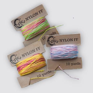 May include: Several spools of nylon thread in various colors, including pink, yellow, and green, are displayed. Each spool is accompanied by a brown tag that reads "SPOOLS NYLON FF" and "10 yards". The thread is tightly wound, showcasing the vibrant color variations.