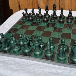 May include: A chess set featuring a square board with alternating dark green and black glittery squares. The set includes a full complement of matte black pieces on one side and metallic dark green pieces on the other, arranged in their starting positions.