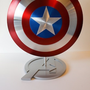 May include: A red, white, and blue Captain America shield replica on a silver stand shaped like the Avengers logo.