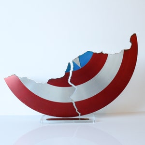 May include: A red, white and blue Captain America shield is broken in half and displayed on a clear acrylic stand. The shield is made of metal and has a distressed look.