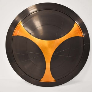 Puede incluir: Un escudo redondo negro con un diseño de estrella de tres puntas en oro en el centro.