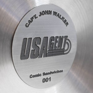 May include: A silver metal disc with the text "CAPT. JOHN WALKER" and "USAGENT" in black lettering. The text "Comic Sandwiches 001" is also printed on the disc.