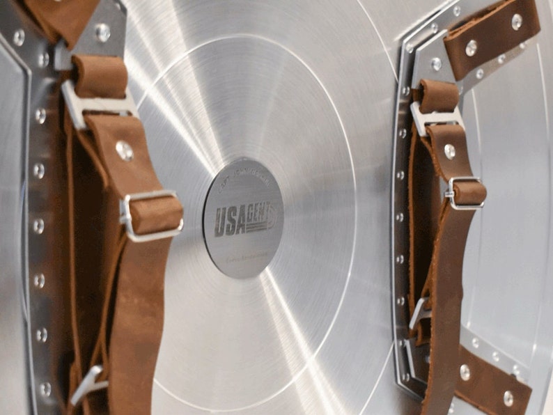 May include: A silver metal shield with brown leather straps. The shield has a circular metal plate in the center with the text "USAGENTS" printed on it.