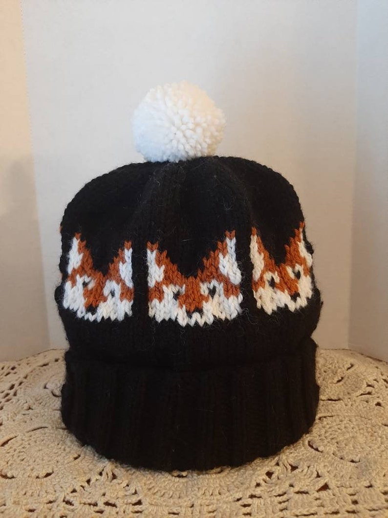 May include: Black knit beanie with a white pom pom and a pattern of three orange foxes on the front.