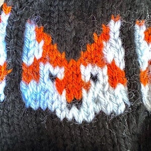 May include: Close-up of a black knitted hat with a white and orange fox design. The fox's face is embroidered with white and orange yarn.