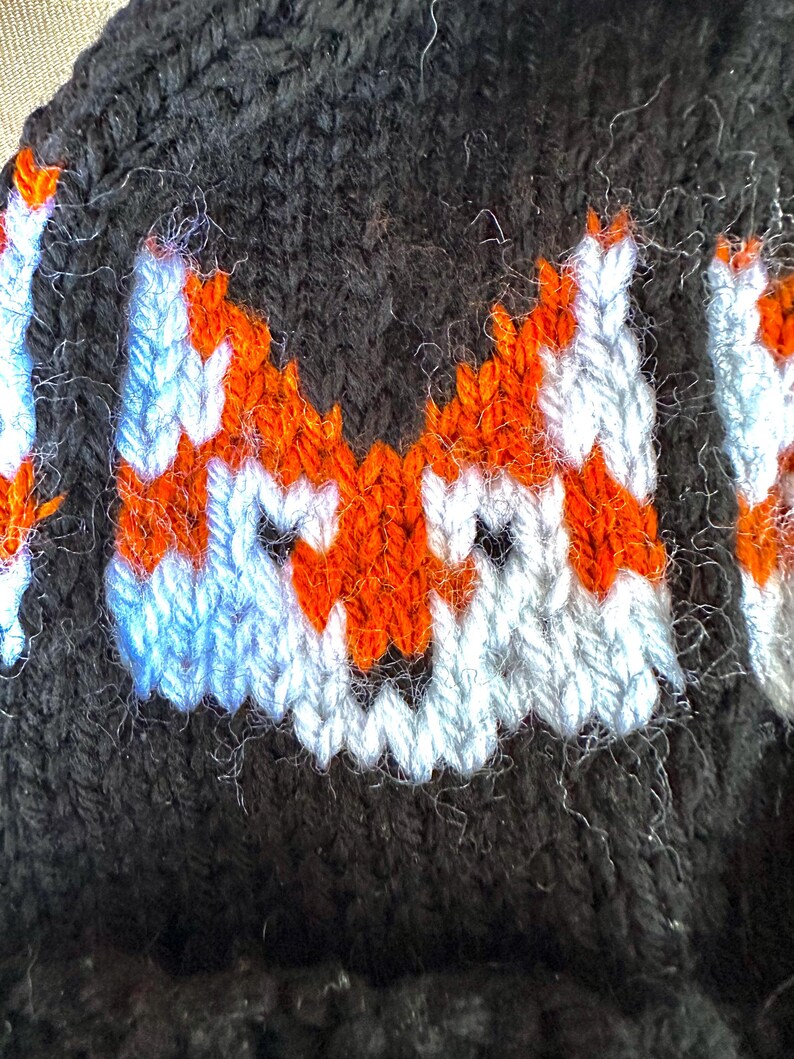 May include: Close-up of a black knitted hat with a white and orange fox design. The fox's face is embroidered with white and orange yarn.
