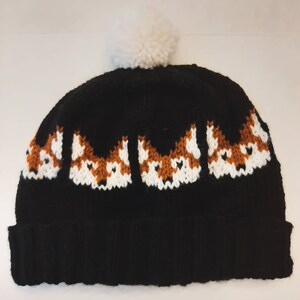 May include: Black knitted beanie with a white pom pom on top. The beanie has a pattern of fox faces in orange, white, and black.
