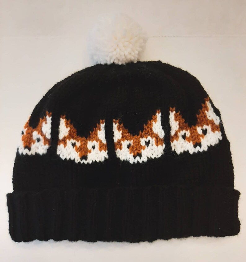 May include: Black knitted beanie with a white pom pom on top. The beanie has a pattern of fox faces in orange, white, and black.