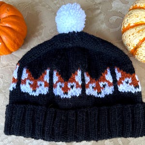May include: Black knit beanie with a white pom pom on top. The beanie has a pattern of orange and white fox faces.