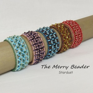 Może przedstawiać: Sześć koralikowych bransoletek w różnych kolorach, w tym turkusowym, fioletowym, niebieskim, brązowym, złotym i czerwonym, ułożonych na brązowej powierzchni. Pod bransoletkami znajduje się napis "The Merry Beader Stardust".