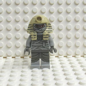 May include: A Lego minifigure of a mummy, featuring a tan headdress and bandages. The figure is primarily gray with detailed wrappings and a stitched mouth. The background is a white baseplate with circular studs.