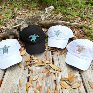 May include: Four baseball caps with a sea turtle design. Two caps are white with a teal turtle design and two caps are white with a multi-colored turtle design. One cap is black with a teal turtle design.
