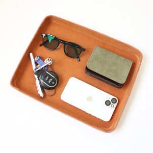 K&ouml;nnte beinhalten: Ein braunes Ledertablett mit einem wei&szlig;en Smartphone, einer gr&uuml;nen Brieftasche, einer schwarzen Sonnenbrille und einem Schl&uuml;sselsatz mit einem Autoschl&uuml;sselanh&auml;nger.