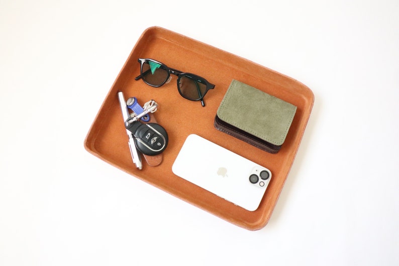 K&ouml;nnte beinhalten: Ein braunes Ledertablett mit einem wei&szlig;en Smartphone, einer gr&uuml;nen Brieftasche, einer schwarzen Sonnenbrille und einem Schl&uuml;sselsatz mit einem Autoschl&uuml;sselanh&auml;nger.