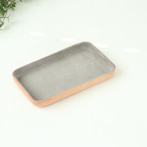 May include: A small, rectangular tray made of leather with a light brown exterior and a gray felt lining.