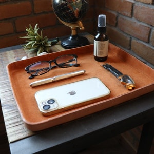 K&ouml;nnte beinhalten: Ein braunes Ledertablett mit einem wei&szlig;en Telefon, einem Stift, einer Brille, einer Uhr und einer Flasche Fl&uuml;ssigkeit auf einem Holztisch.