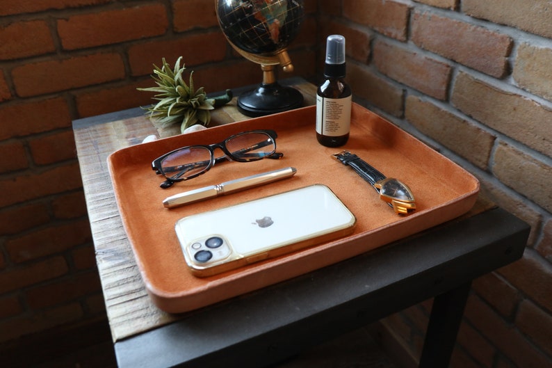 K&ouml;nnte beinhalten: Ein braunes Ledertablett mit einem wei&szlig;en Telefon, einem Stift, einer Brille, einer Uhr und einer Flasche Fl&uuml;ssigkeit auf einem Holztisch.