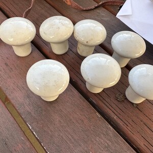 May include: Seven off-white ceramic mushroom-shaped cabinet knobs. The knobs have a speckled appearance and a rounded top with a short, cylindrical base. They are arranged on a weathered, reddish-brown wooden surface.