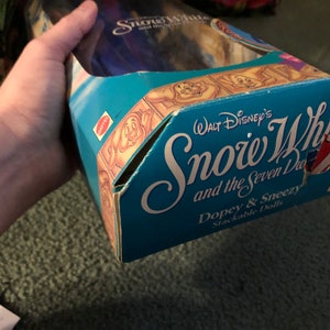 Puede incluir: Una caja azul y blanca para un conjunto de mu&ntilde;ecas apilables con Blancanieves y los Siete Enanitos. La caja tiene una imagen de Tont&iacute;n y Estornudo en la parte delantera y el texto "WALT Disney's Snow White and the Seven Dwarfs Dopey & Sneezy Stackable Dolls".