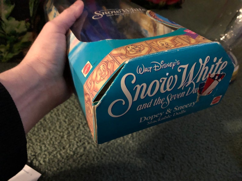 Puede incluir: Una caja azul y blanca para un conjunto de mu&ntilde;ecas apilables con Blancanieves y los Siete Enanitos. La caja tiene una imagen de Tont&iacute;n y Estornudo en la parte delantera y el texto "WALT Disney's Snow White and the Seven Dwarfs Dopey & Sneezy Stackable Dolls".