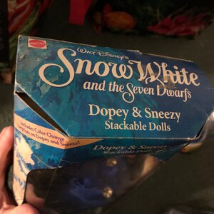 Puede incluir: Una caja azul y blanca para un conjunto de mu&ntilde;ecas apilables con Dopey y Sneezy de la pel&iacute;cula de Disney Blancanieves y los Siete Enanitos. La caja incluye el texto "Blancanieves y los Siete Enanitos" y "Dopey & Sneezy Stackable Dolls".