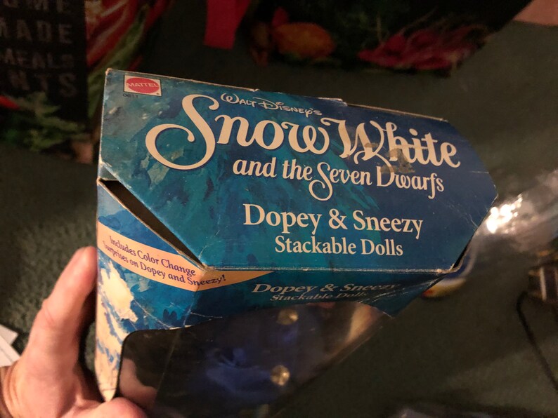 Puede incluir: Una caja azul y blanca para un conjunto de mu&ntilde;ecas apilables con Dopey y Sneezy de la pel&iacute;cula de Disney Blancanieves y los Siete Enanitos. La caja incluye el texto "Blancanieves y los Siete Enanitos" y "Dopey & Sneezy Stackable Dolls".