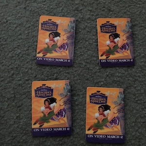May include: Four promotional cards for Disney's "The Hunchback of Notre Dame" on a gray surface. Each card features the movie's title and artwork of the characters. The text "ON VIDEO MARCH 4!" is displayed on each card.