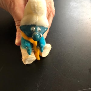May include: A blue Smurf figurine wearing a white hat and a yellow scarf. The Smurf is sitting with its legs crossed and has a white and blue outfit.