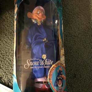 Puede incluir: Un juego de dos mu&ntilde;ecas apilables de Dopey y Sneezy de Blancanieves y los Siete Enanitos de Disney. Las mu&ntilde;ecas est&aacute;n vestidas con sus ic&oacute;nicos atuendos azules y morados. La caja presenta el logotipo de Disney y el t&iacute;tulo "Blancanieves y los Siete Enanitos".