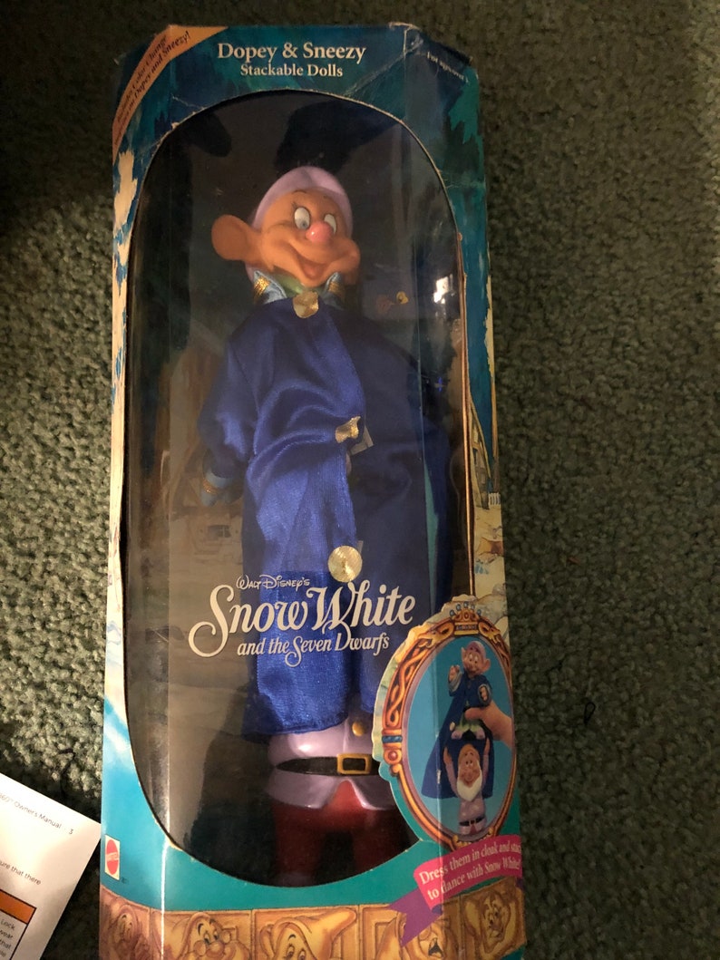 Puede incluir: Un juego de dos mu&ntilde;ecas apilables de Dopey y Sneezy de Blancanieves y los Siete Enanitos de Disney. Las mu&ntilde;ecas est&aacute;n vestidas con sus ic&oacute;nicos atuendos azules y morados. La caja presenta el logotipo de Disney y el t&iacute;tulo "Blancanieves y los Siete Enanitos".