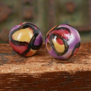 May include: Two round ceramic beads with a purple, yellow, red, and black abstract pattern.