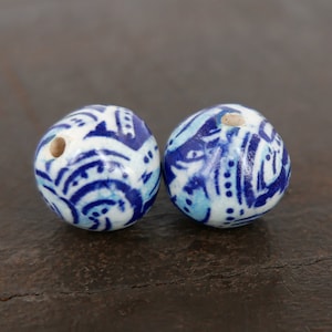 May include: Two round ceramic beads with a white base and intricate blue patterns. The beads have a hole for stringing and feature a mix of dark and light blue designs, creating a decorative element.